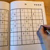 Sudoku puzzle book with a partially completed grid and a hand holding a pen.