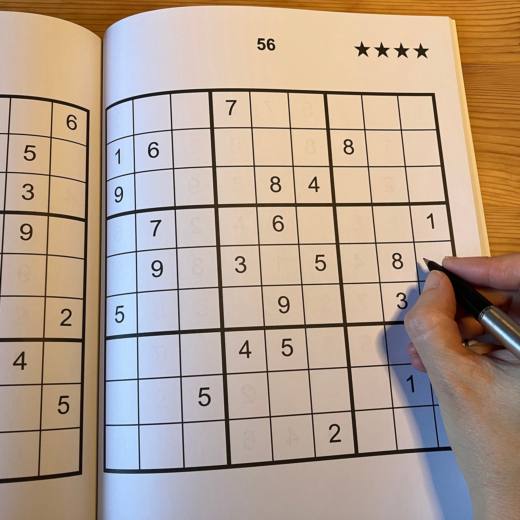 Sudoku puzzle book with a partially completed grid and a hand holding a pen.