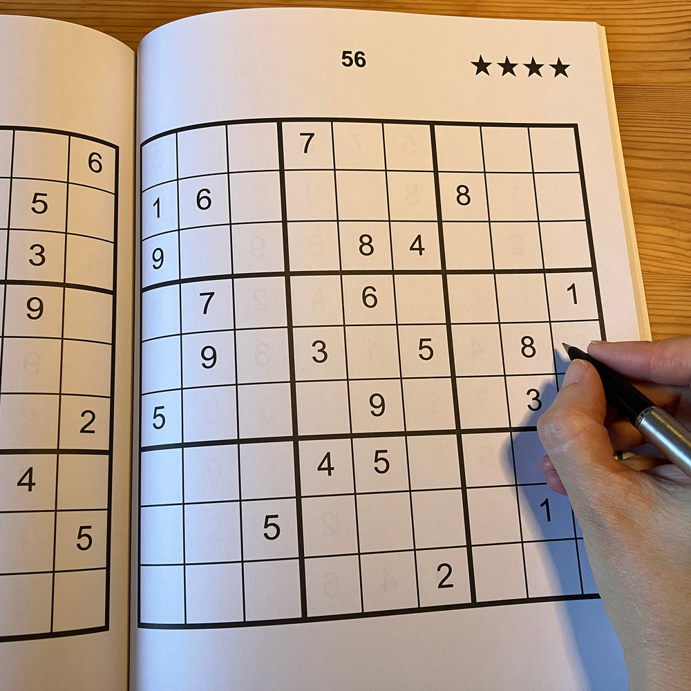 Sudoku puzzle book with a partially completed grid and a hand holding a pen.