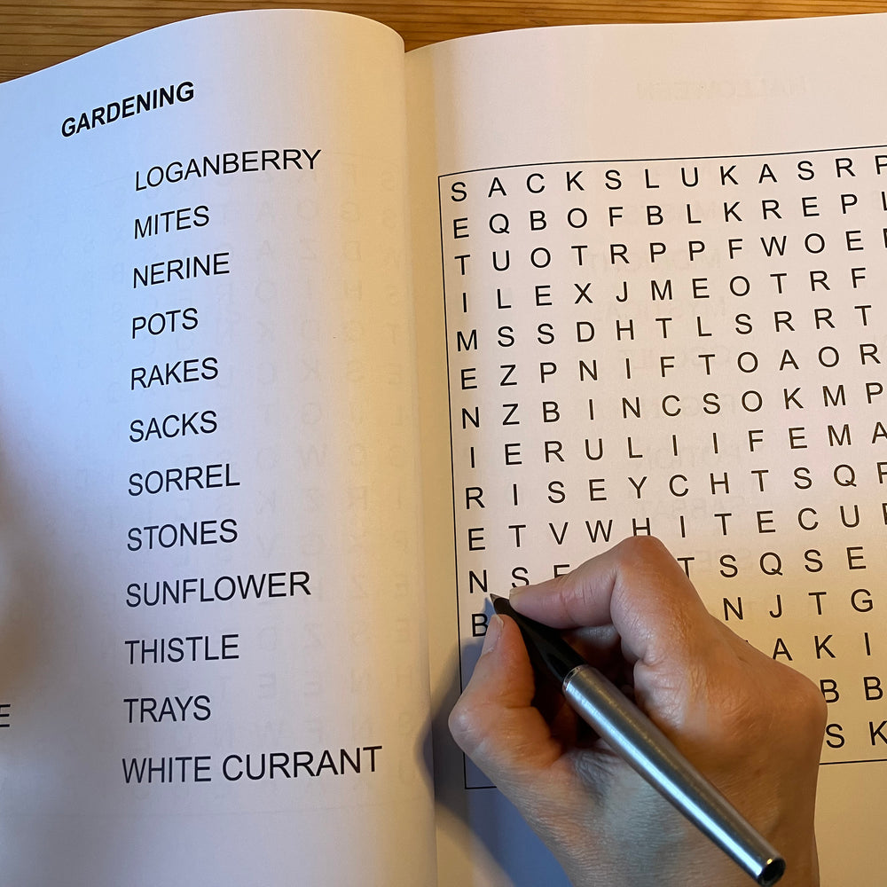 Hand holding a pen over an open book with a word search and gardening-related words listed.