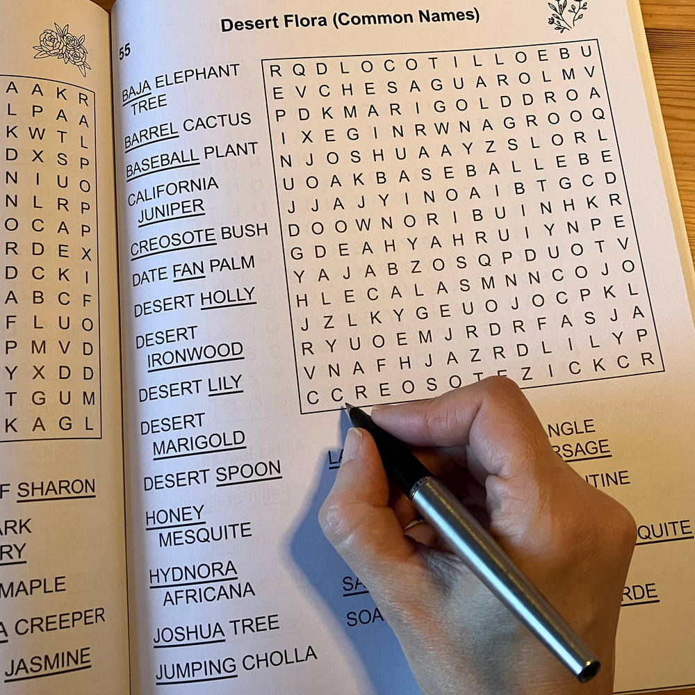 Hand holding a pen over a book with a word search puzzle on desert flora.