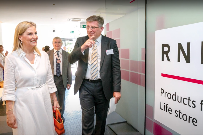 Her Royal Highness, The Duchess of Edinburgh, Global Ambassador for the International Agency for the Prevention of Blindness (IAPB), a white woman with shoulder length blonde hair, wearing a white shirt dress and carrying an orange handbag is being shown a the RNIB Products for life store by the Chief Executive of the RNIB, Matt Stringer, a white man with short blond grey hair wearing glasses, a grey suit with a white shirt and a yellow, blue and white diagonally striped tie.