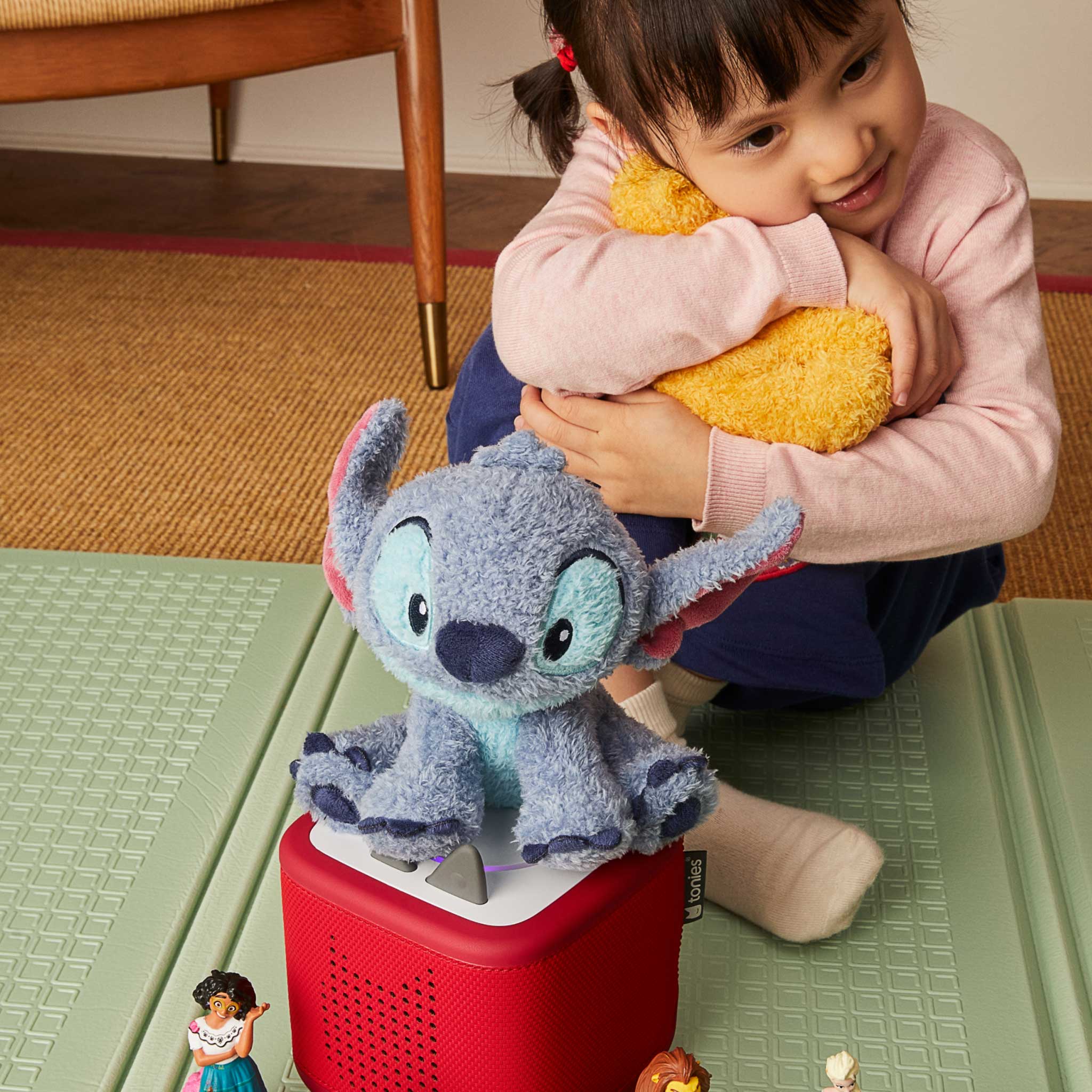 Child holding a yellow plush toy with a gray plush toy resembling Stitch on a red box.