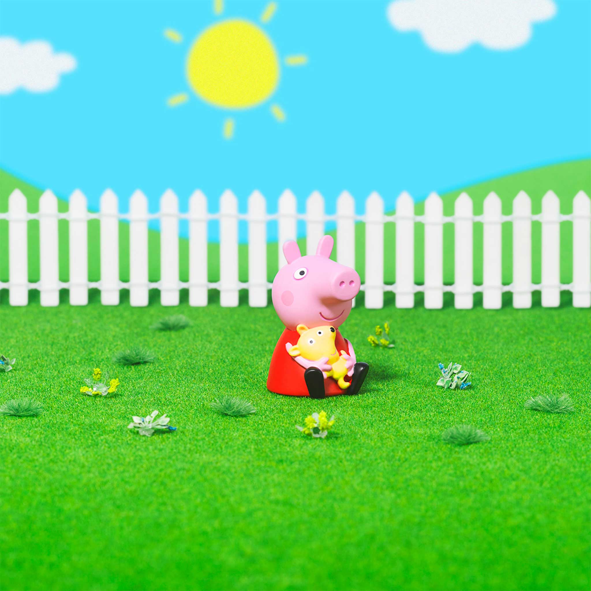 Toy figure of a pink character holding a yellow figure on a grassy area with a white picket fence and blue sky.