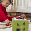 Child playing with toy cars near a green electronic device on a wooden surface.