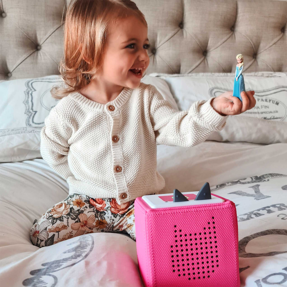 Child holding a toy figure next to a pink toy box on a bed