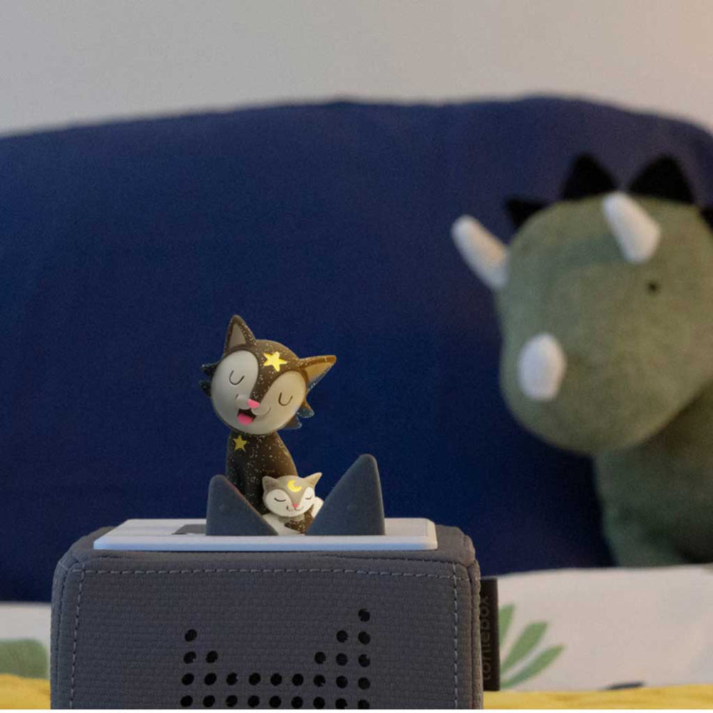 Small cat figurine on a gray electronic device with a blue cushion and green plush toy in the background.