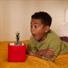 A young boy in a green sweater lies on a yellow quilted bed, looking surprised at a red children's speaker with a Jack Skellington figure on top.