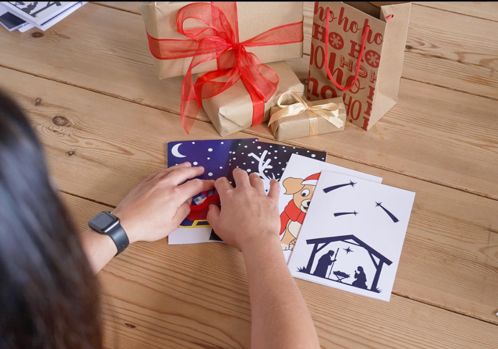 A woman feels the tactile images on inclusive Christmas cards on a wooden table. Nearby are gift-wrapped presents and a gift bag