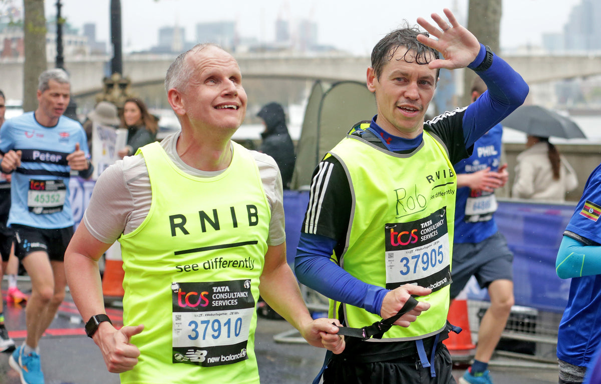 Two white men in day-glo yellow vests taking part in the London marathon. The runner on the left is blind and holding a running tether which is held at the other end by his guide runner. The guide is waving to someone.