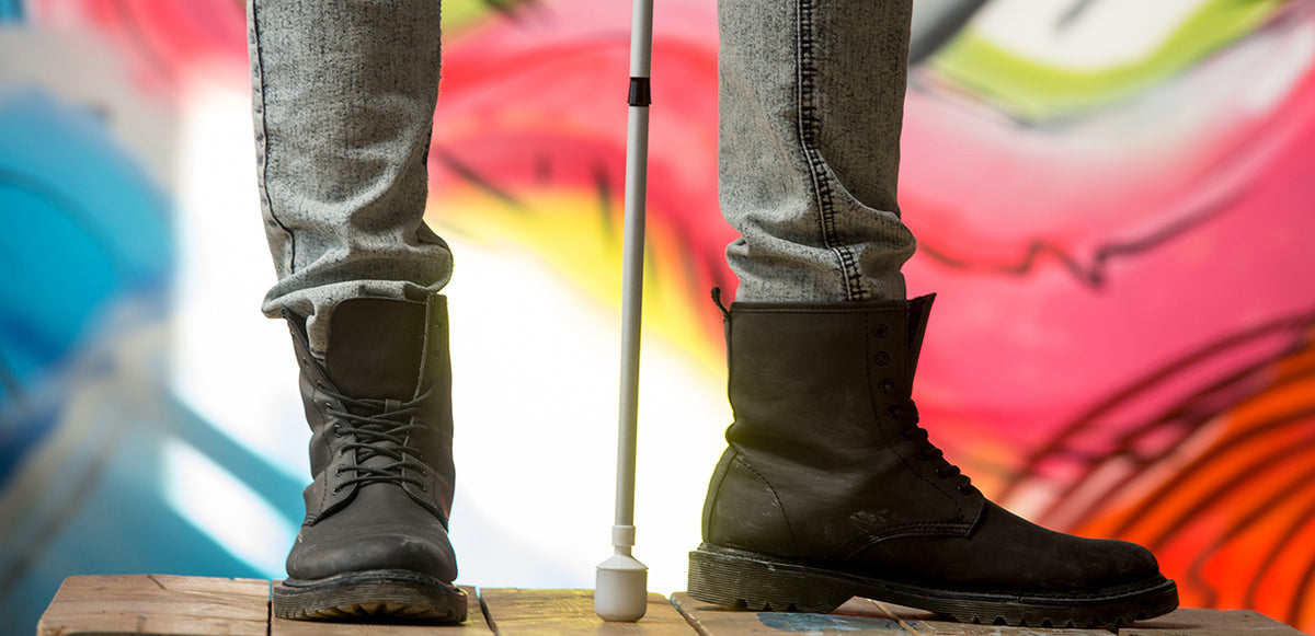 A person's booted feet standing in front of colourful graffiti with a white cane with roller marshmallow tip being held between their feet. 