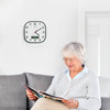 An elderly woman with gray hair and glasses sits on a gray sofa reading a magazine, with a black-framed round wall clock on the white wall behind her.
