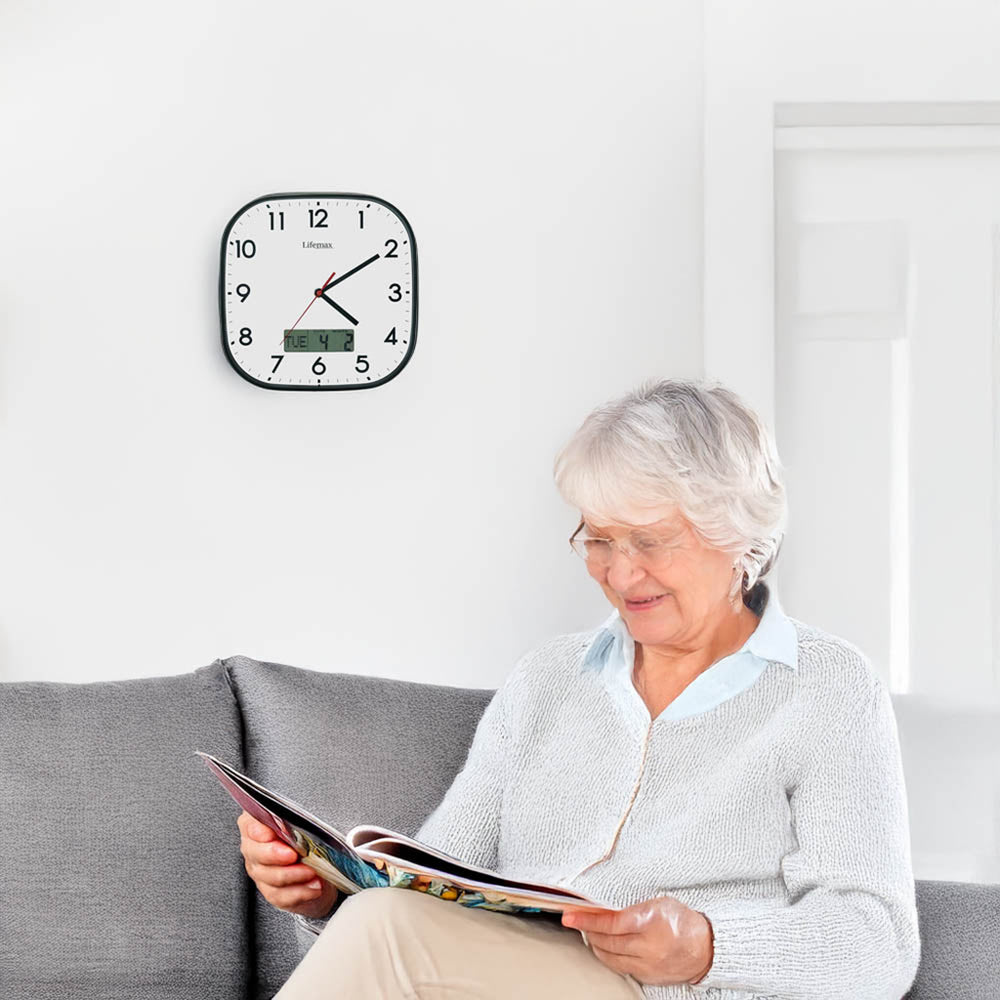 An elderly woman with gray hair and glasses sits on a gray sofa reading a magazine, with a black-framed round wall clock on the white wall behind her.