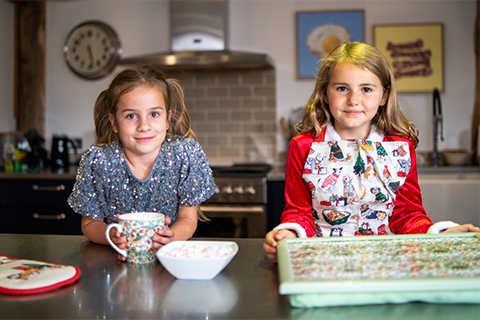 Two girls in festive clothing lean on a kitchen counter. One holds a William Morris mug, the other wears a Merry Mutts apron and holds a William Morris laptray