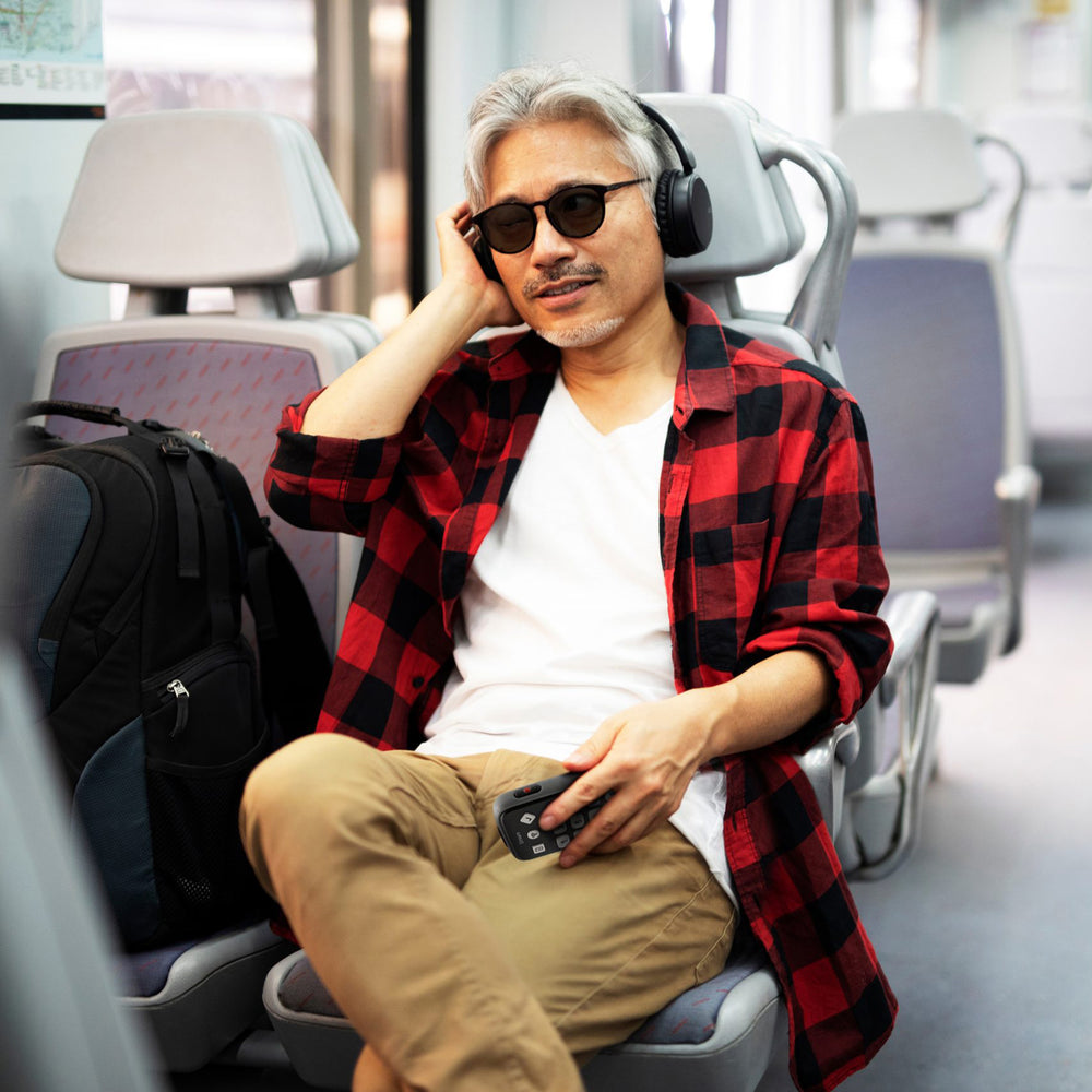 A man with white hair and sunglasses sitting on a train listening to a Victor Stream 3 through wireless headphones