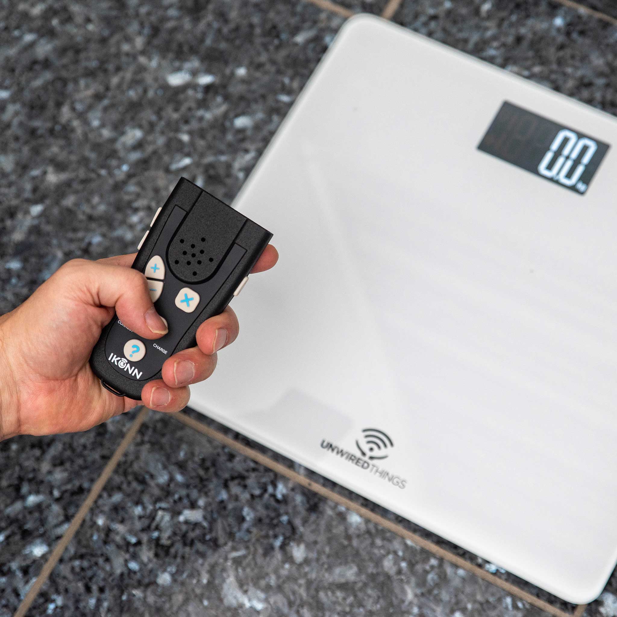 Hand holding the IKONN control device next to the IKONN digital scale on a textured surface