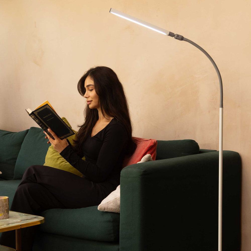 A woman sits on a dark green sofa reading a book in a cosy living room, with a tall modern floor lamp casting light next to her.