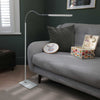 A modern white floor lamp with a flexible gooseneck arm and rectangular head, standing on a patterned rug next to a grey fabric sofa in a dark green room.