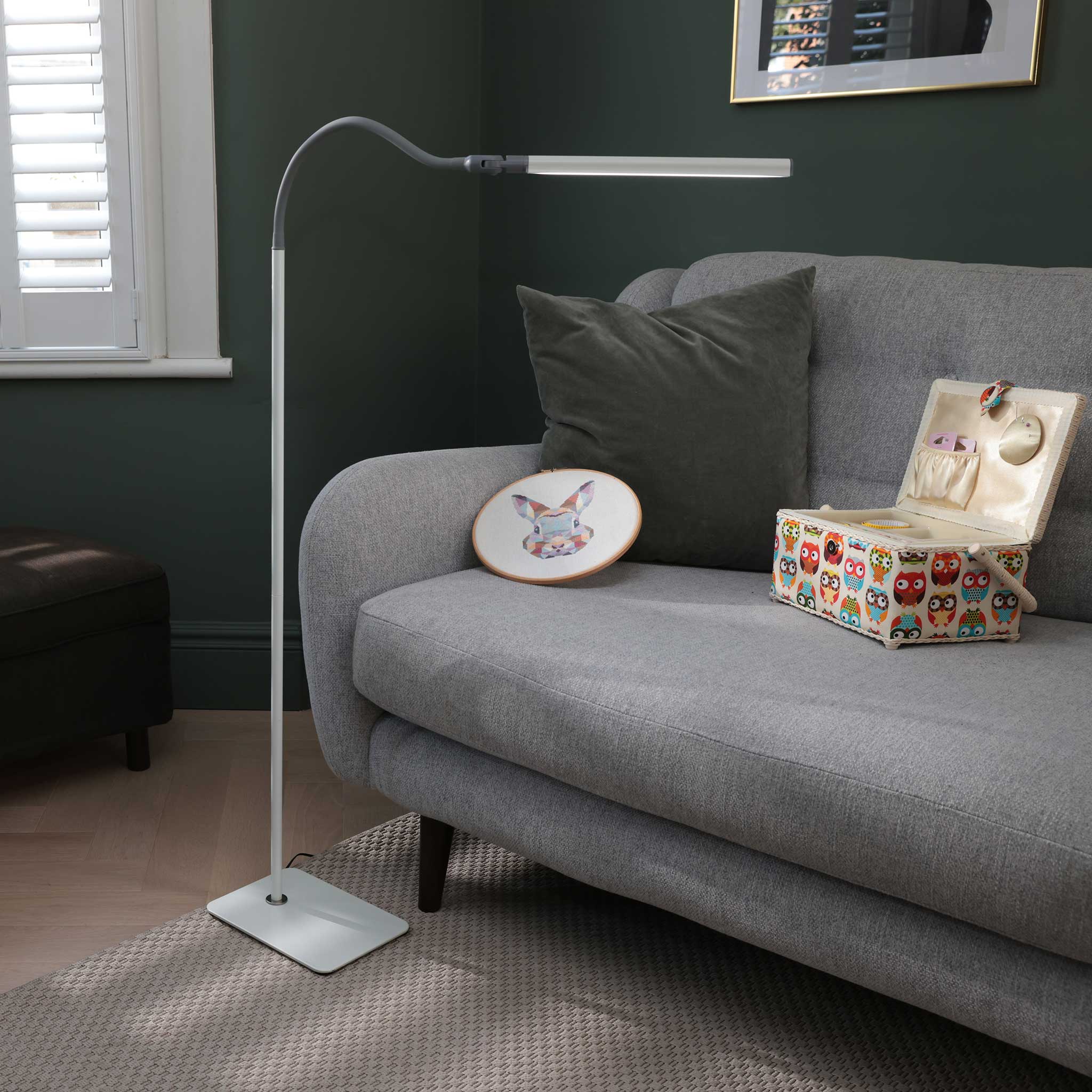 A modern white floor lamp with a flexible gooseneck arm and rectangular head, standing on a patterned rug next to a grey fabric sofa in a dark green room.