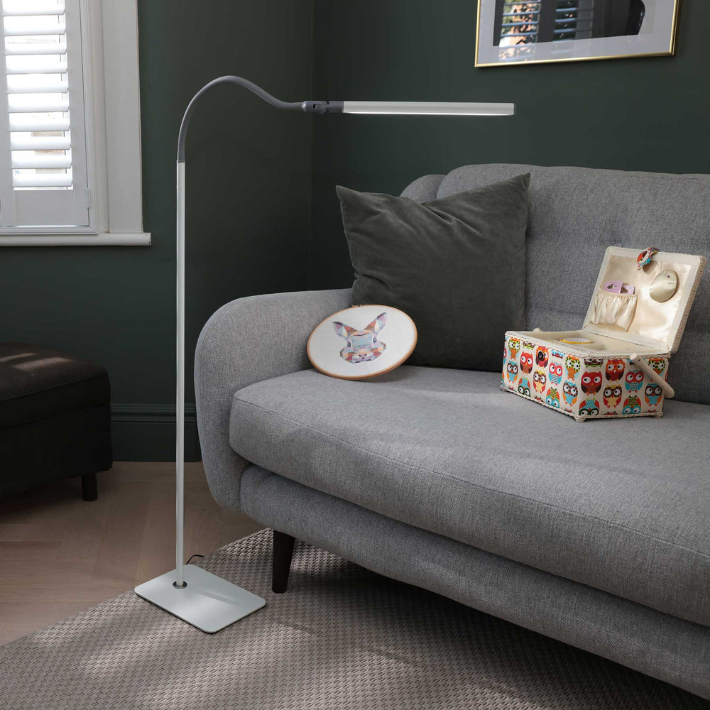 A modern white floor lamp with a flexible gooseneck arm and rectangular head, standing on a patterned rug next to a grey fabric sofa in a dark green room.