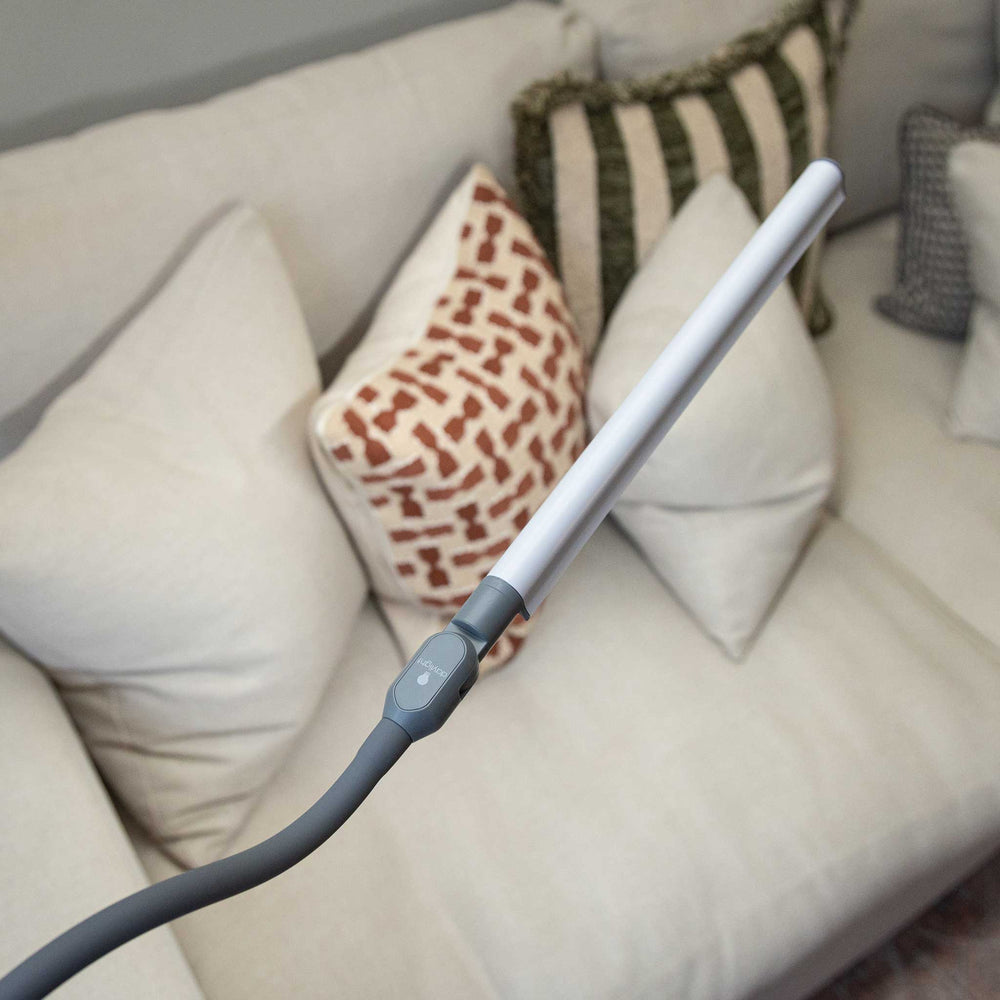 A modern LED lamp with a long white rectangular light bar and a flexible grey neck, shown with a beige sofa with decorative pillows in the background.