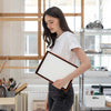 A woman in a white t-shirt and dark jeans holds the A4 board with a black and orange border in a bright, modern art studio.