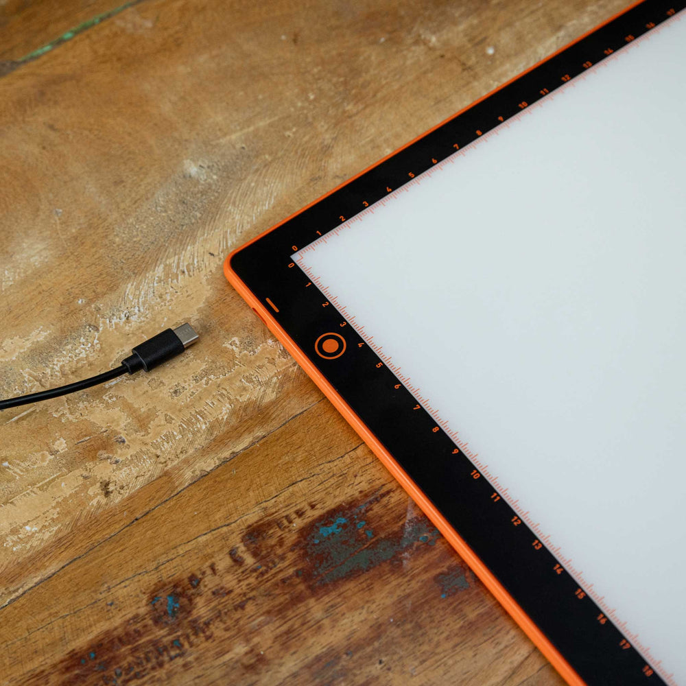 A close-up of a rectangular LED light panel with a bright white illuminated surface and an orange border, placed on a rustic wooden table. A black USB charging cable is visible on the left side.