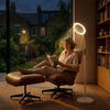 A woman relaxes in a brown leather lounge chair with an ottoman beside a glowing white floor lamp in a cozy room. Large windows behind her show a nighttime garden view, and a bookshelf stands to the right.