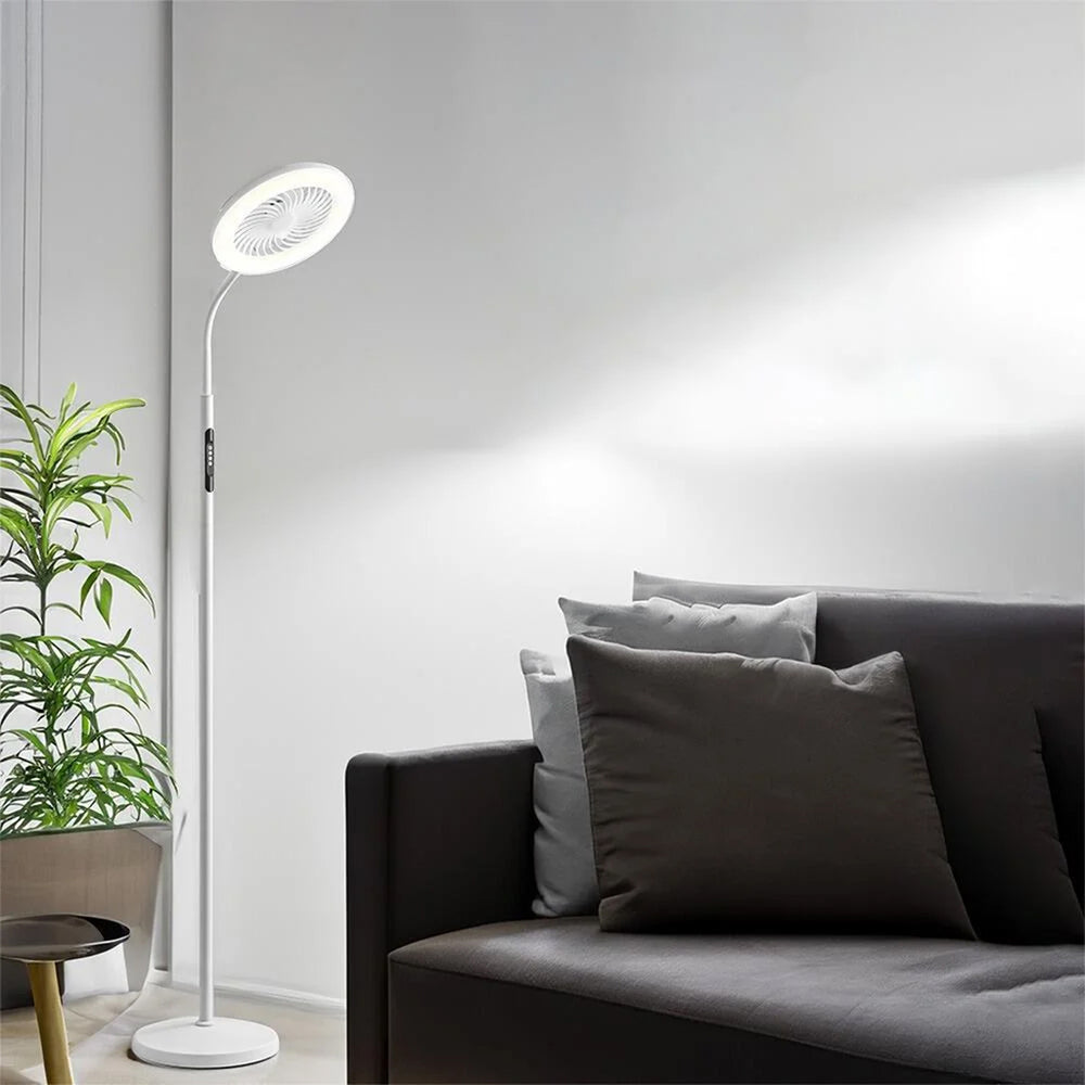 A modern white floor lamp with a circular head and integrated fan in the corner of a well lit room. The lamp has a plant behind it and a dark grey sofa next to it.