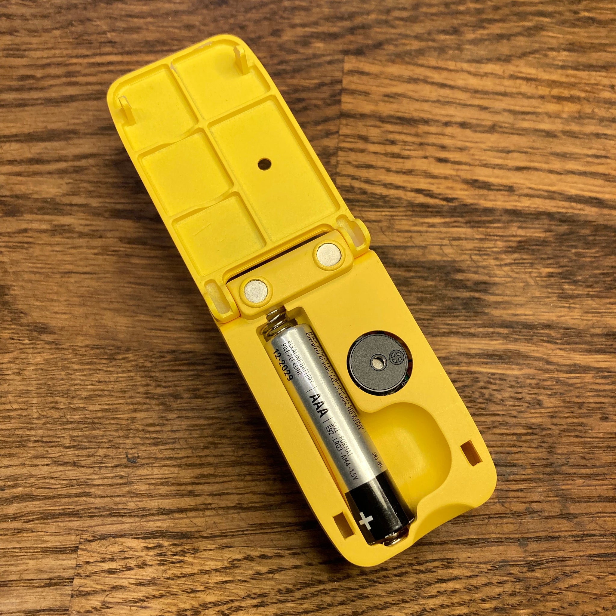 Liquid level indicator with battery compartment open with a AAA battery inside, this is on a wooden surface.