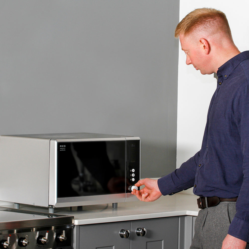 The RNIB talking microwave on a counter top in a kitchen setting A man wearing a dark blue shirt and grey trousers is using the controls to set the timer.
