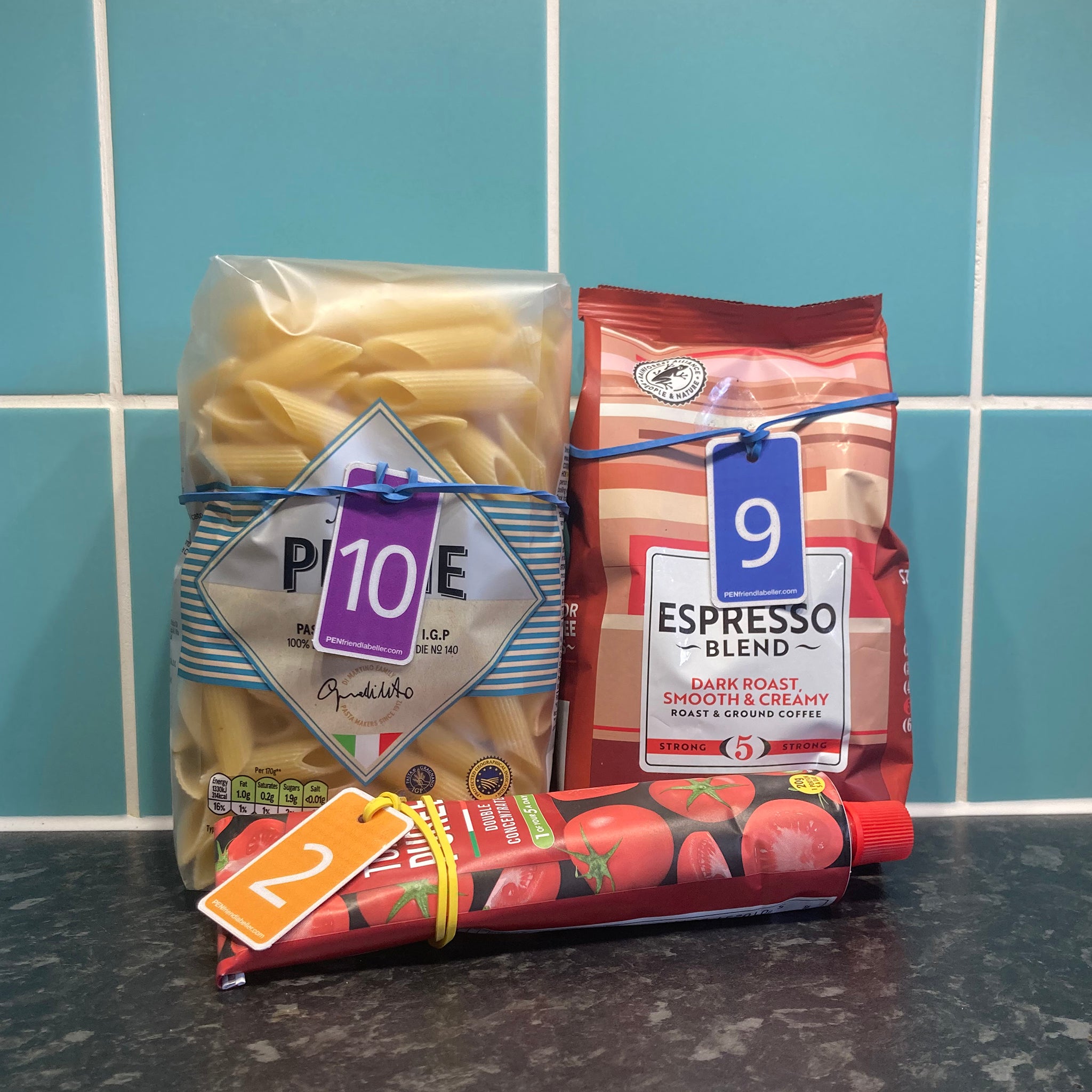 Rubber band fobs attached to groceries (bag of pasta, bag of ground coffee, tube of tomato paste) on a kitchen worktop