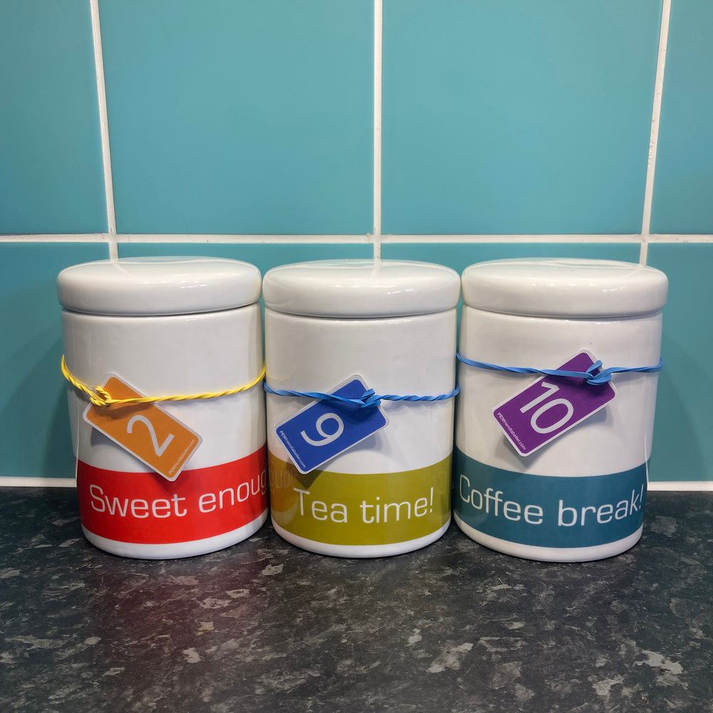 Three ceramic caddies for sugar, tea and coffee on a worktop, each labelled with a rubber band fob