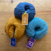 Three balls of yarn on a wooden surface, each one labelled with a rubber band fob.