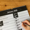 Calendar for January 2026 on a wooden surface with a hand holding a pen.