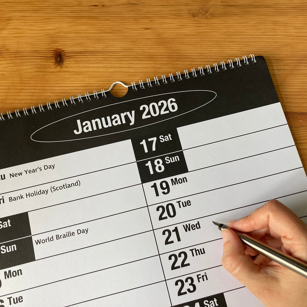 Calendar for January 2026 on a wooden surface with a hand holding a pen.