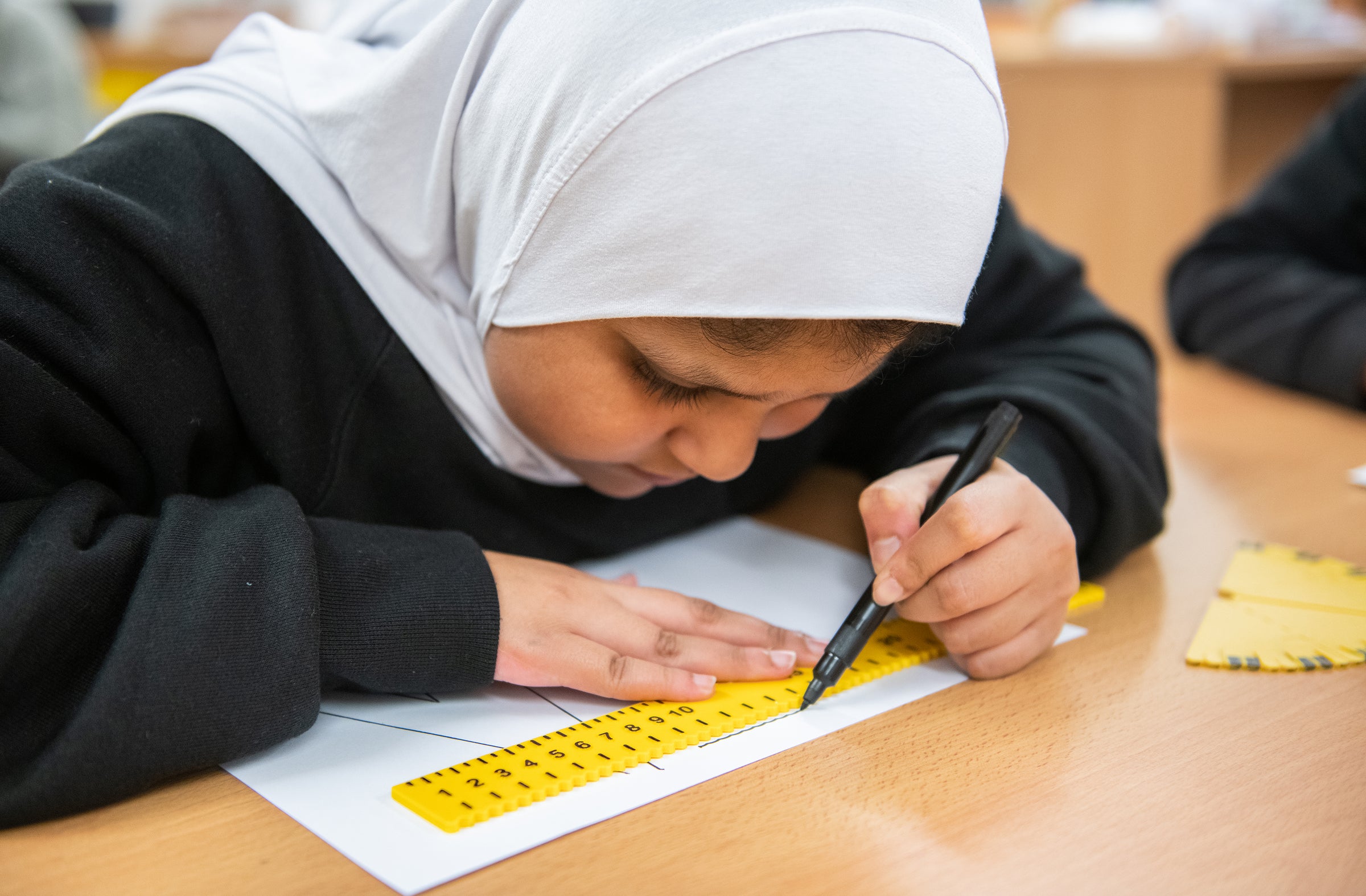 A young girl in black pullover and white hijab uses a thick black pen and RNIB's tactile ruler to draw a line