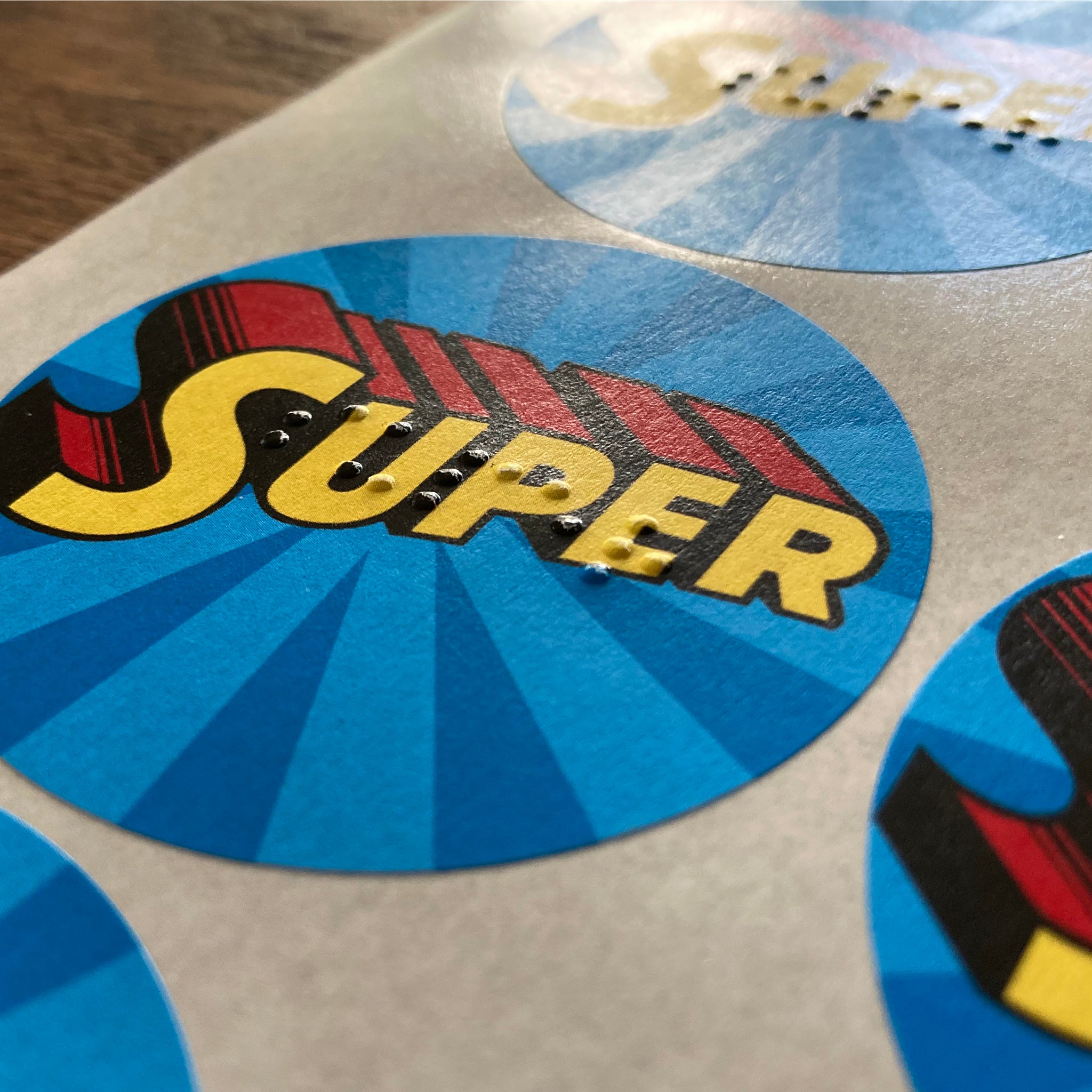 Circular stickers with superhero-themed design with braille.