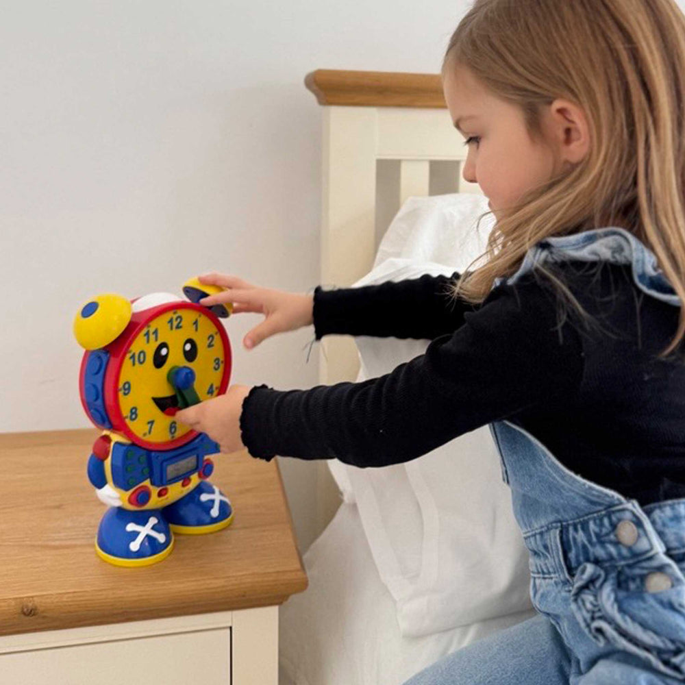 A young child in denim overalls and a black long-sleeve top is touching a colorful toy robot clock on a wooden bedside table next to a bed.