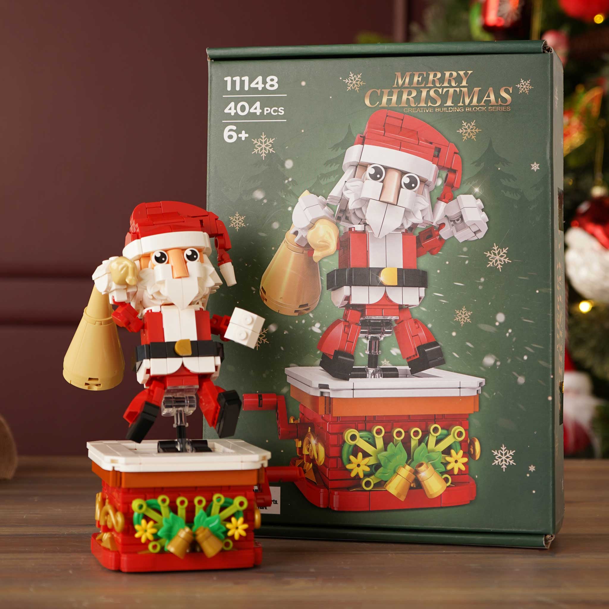 A Christmas Santa Claus building block set shown with its retail box on a wooden surface. The assembled toy Santa stands on a red and white base holding a sack, with a matching box behind it featuring festive graphics and the text 'MERRY CHRISTMAS' and '404 pieces'.