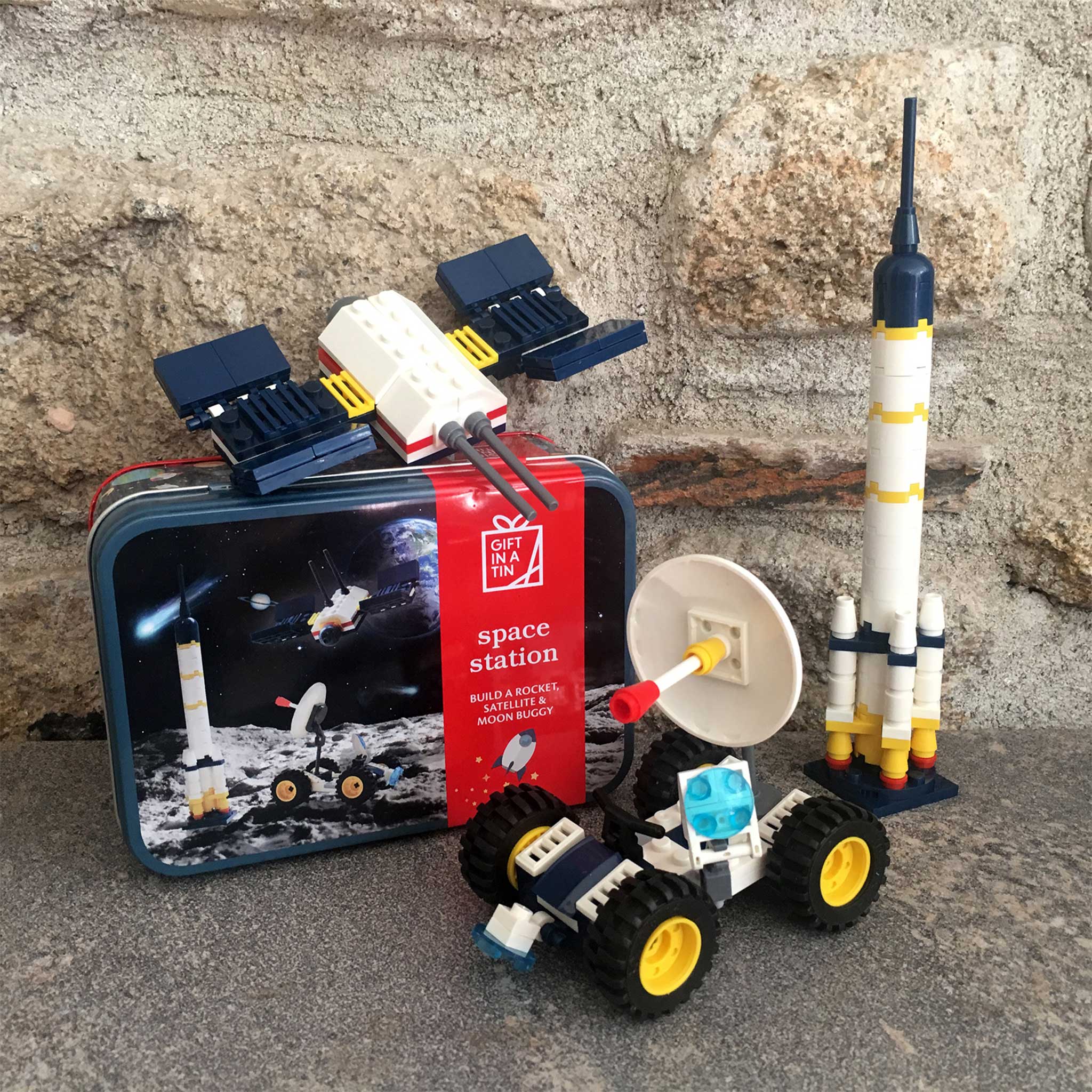 A toy construction set featuring a space station model, a rocket, a rover, and a satellite dish, displayed in front of its illustrated tin packaging on a stone surface.