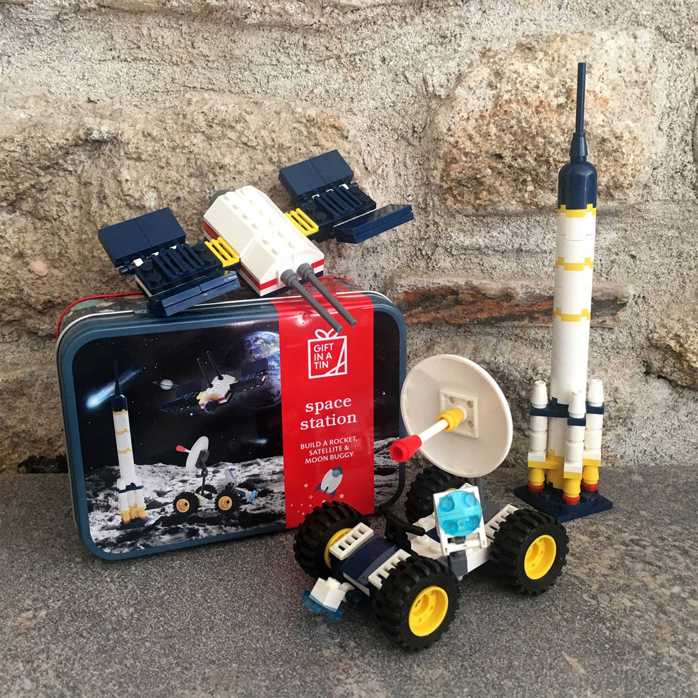 A toy construction set featuring a space station model, a rocket, a rover, and a satellite dish, displayed in front of its illustrated tin packaging on a stone surface.
