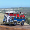 A metal tin bus toy with a silver chrome body, red interior, and blue side panel, displayed on a wooden surface with a blurred outdoor landscape background.