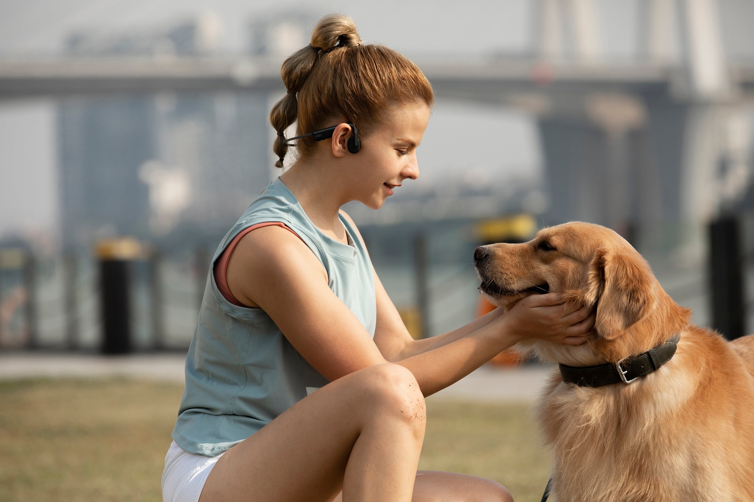 A young white woman wearing bone-conduction headphones and sportswear pets a golden retriever's cheeks.