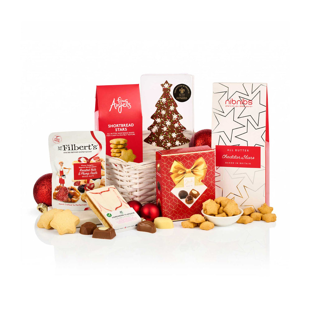 A Christmas-themed gourmet gift hamper featuring an assortment of chocolates, biscuits, and treats arranged around a white wicker basket on a white background. 