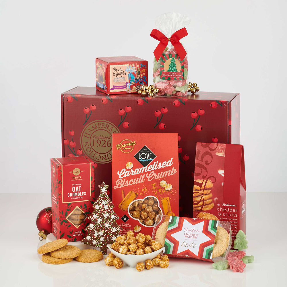 A holiday gift hamper set from Malmaison featuring a large red Christmas-themed box, smaller red gift boxes, a bag of ornament-shaped chocolates tied with a red ribbon, a box of caramelised biscuit crumb, oat cookies, popcorn, and a small decorative tree, arranged on a white background.