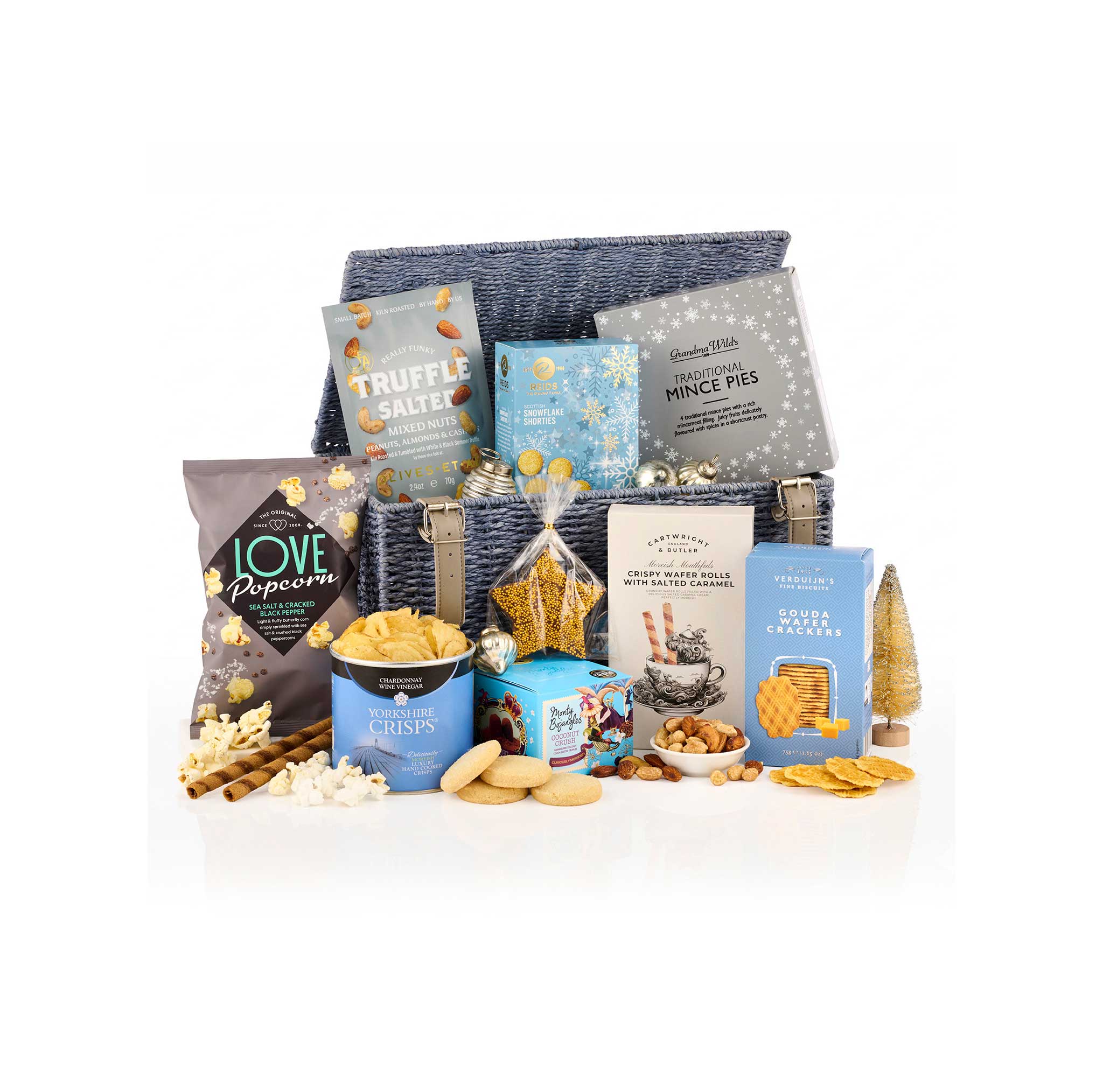 A gourmet gift hamper filled with assorted snacks, biscuits, and packaged treats arranged in front of a dark blue woven basket on a white background. Visible items include Love Popcorn bags, Truffle Salt, Mince Pies, Butter Cookies, and other snack boxes.