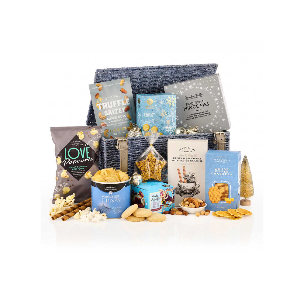 A gourmet gift hamper filled with assorted snacks, biscuits, and packaged treats arranged in front of a dark blue woven basket on a white background. Visible items include Love Popcorn bags, Truffle Salt, Mince Pies, Butter Cookies, and other snack boxes.