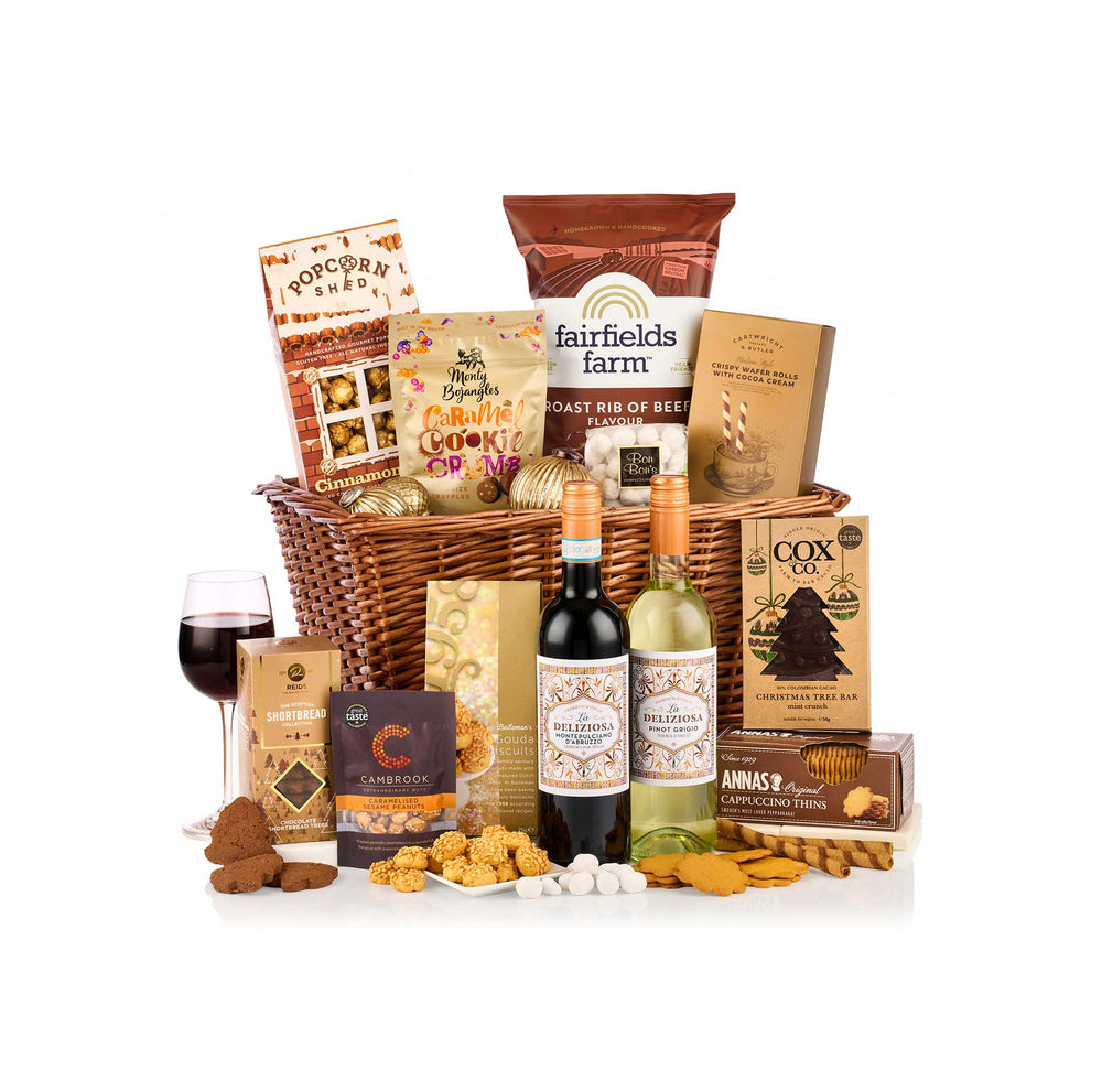 A gourmet gift hamper arranged in a woven wicker basket on a white background, featuring two wine bottles, a glass of red wine, cookies, biscuits, and packaged snacks from brands including Fairfields Farm, Cox's, and Sensory Studies.