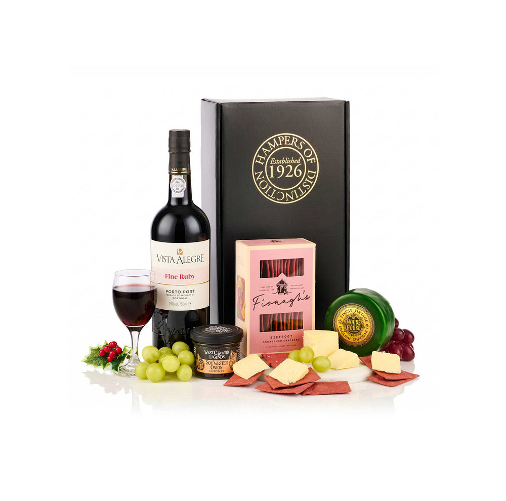 A curated gift set featuring a bottle of Vista Alegre Fine Bodegaria Port wine, a glass of red wine, a black box with gold lettering, a pink box labeled Bineaghs, a green and gold cheese wheel, a small round cheese container, and assorted cheeses and cured meats arranged on a white background with grapes and red berries as props.